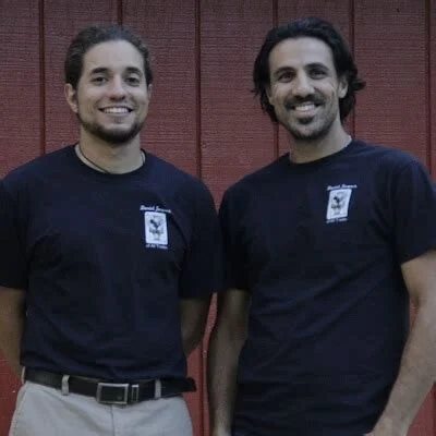 Two members of the Daniel Jacques of All Trades team in navy company T-shirts, smiling in front of a red wall